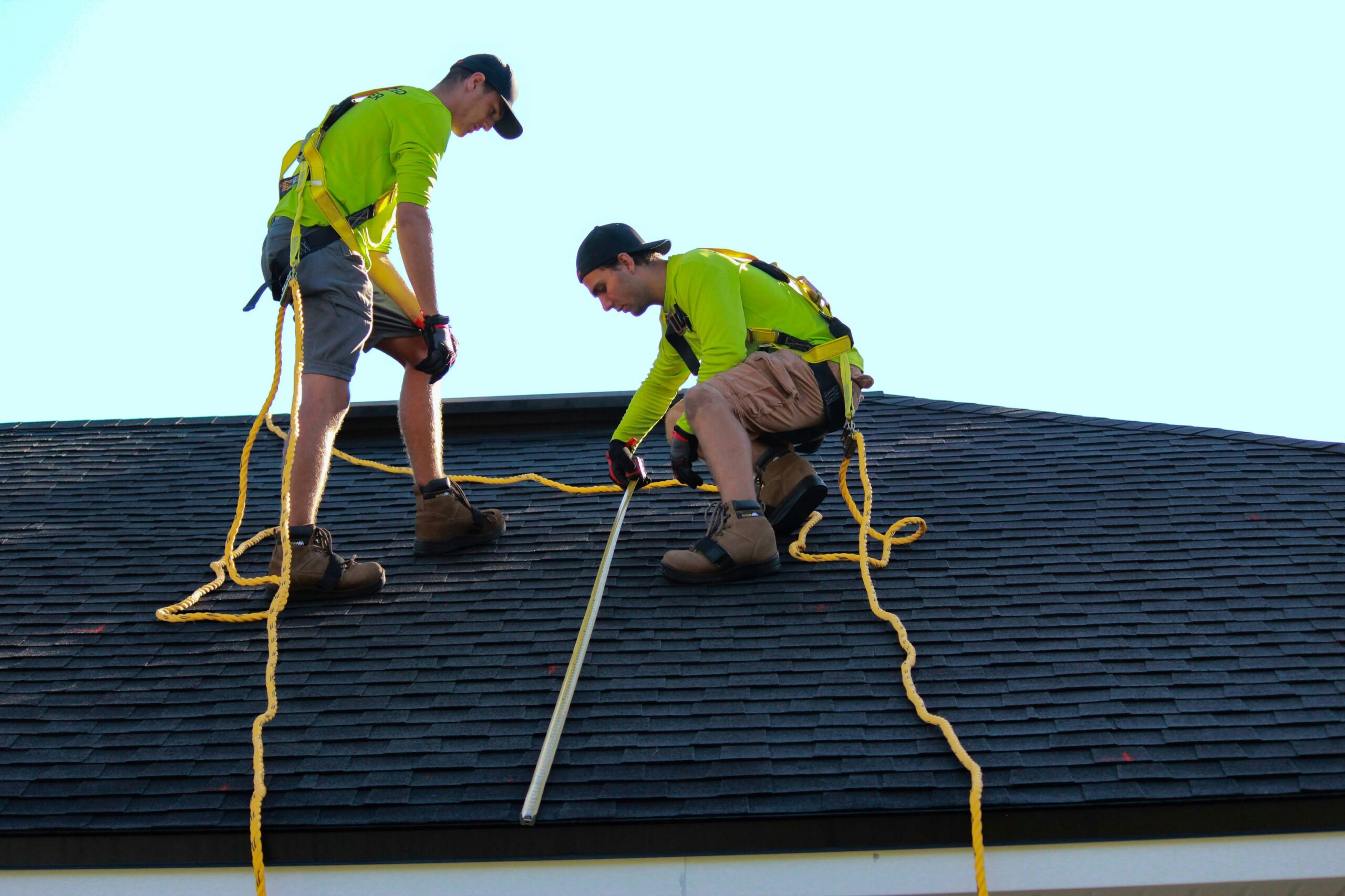 Two roofers wearing safety harnesses measuring and inspecting a residential roof for repair or replacement under clear skies. | Roofing in Georgetown, TX